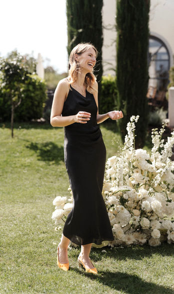 Annemarie Fritsche steht lachend im schwarzen Kleid neben weißen Blumenarrangements im Garten einer Villa.
