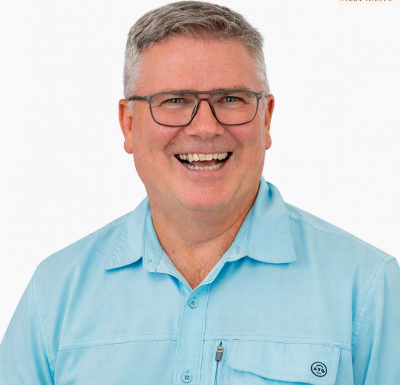 Square-ish portrait headshot of a smiling middle-aged man in a light blue shirt on a clean white background, centered, with shoulders visible.