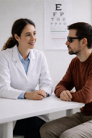 An optometrist sits across from a blind man wearing dark glasses in a clean clinical setting, engaging in a calm, attentive conversation. With an eye chart in the background, the scene reflects a supportive vision care consultation focused on understanding and patient-centered care.
