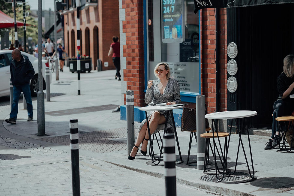 Reiki Woman Wellness - Woman in a gray dress sips coffee at an outdoor cafe table on a city street. A brick wall and passerby in the background. Sign reads "Shop 24/7."