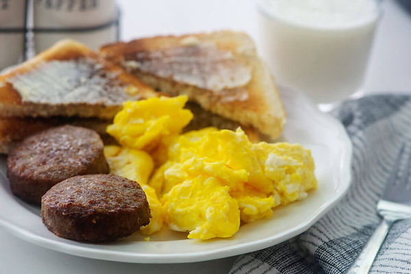 A close-up, eye-level shot of a breakfast plate featuring two sausage patties, a generous portion of fluffy scrambled eggs, and two slices of buttered toast cut into triangles. A glass of milk is blurred in the background on the right