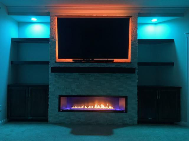 TV above a linear fireplace with dark cabinets and teal accent lighting.