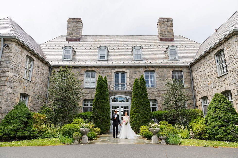 NYC wedding DJ Bride and groom stand hand in hand outside a stone mansion, surrounded by manicured greenery and tall shrubs on an overcast day