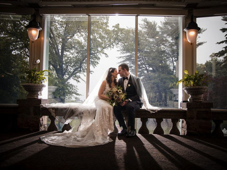 nyc wedding dj Bride and groom sit on a stone bench by large windows, smiling and touching foreheads, with bouquet, veil, and trees outside.