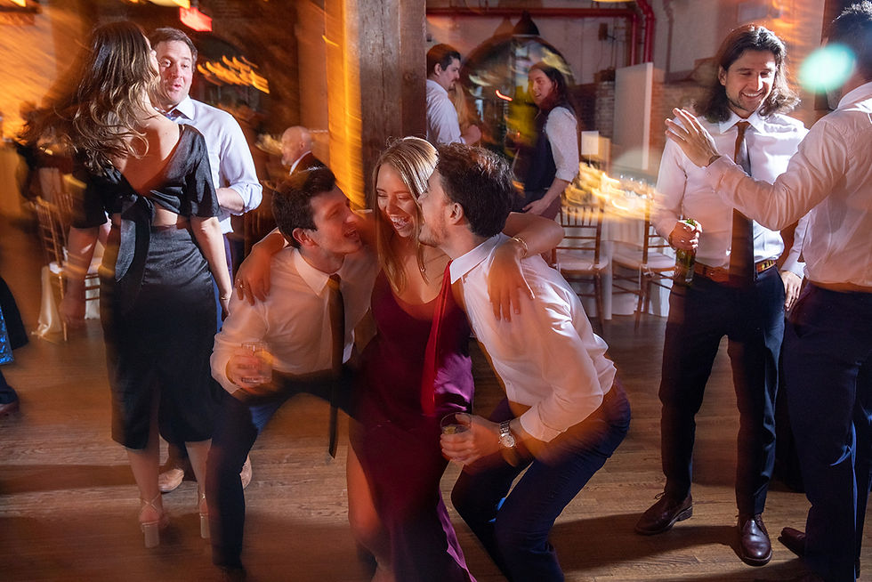 NYC People dance joyfully at a party, with warm lighting and blurred motion around them. A woman in a burgundy dress smiles, surrounded by friends.