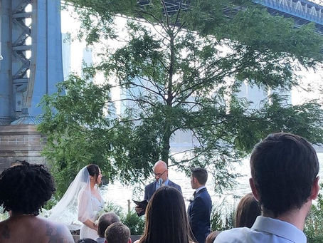 nyc wedding dj Outdoor wedding ceremony by the river under a bridge, bride and groom with officiant before seated guests.