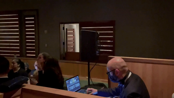 nyc wedding dj new york wedding timeline Man in a suit works on a laptop beside a monitor in a dim courtroom-like room while seated people watch.