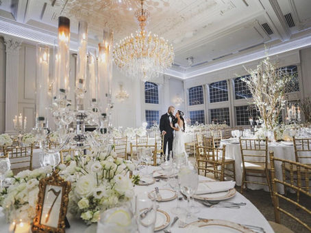 nyc wedding dj bergen county dj Bride and groom embrace in a grand white ballroom with crystal chandeliers, candlelit tables, white flowers, and gold chairs.