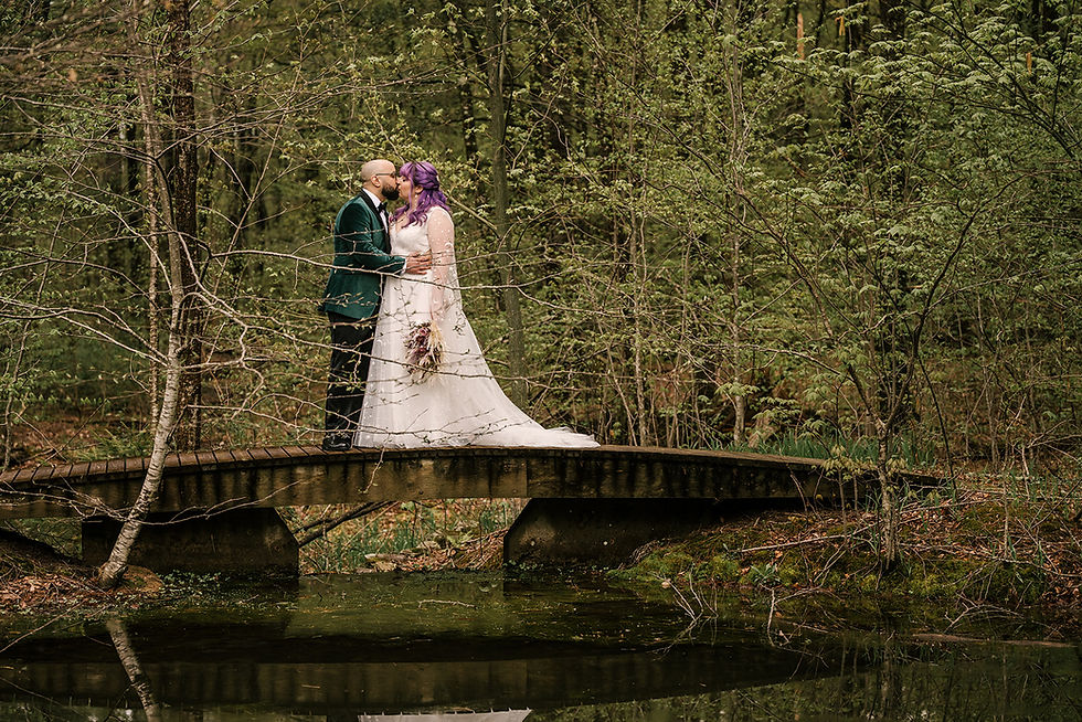 nyc ulster county wedding dj Wedding couple kissing on a wooden bridge in a lush forest, bride in white dress with purple hair, groom in green suit.