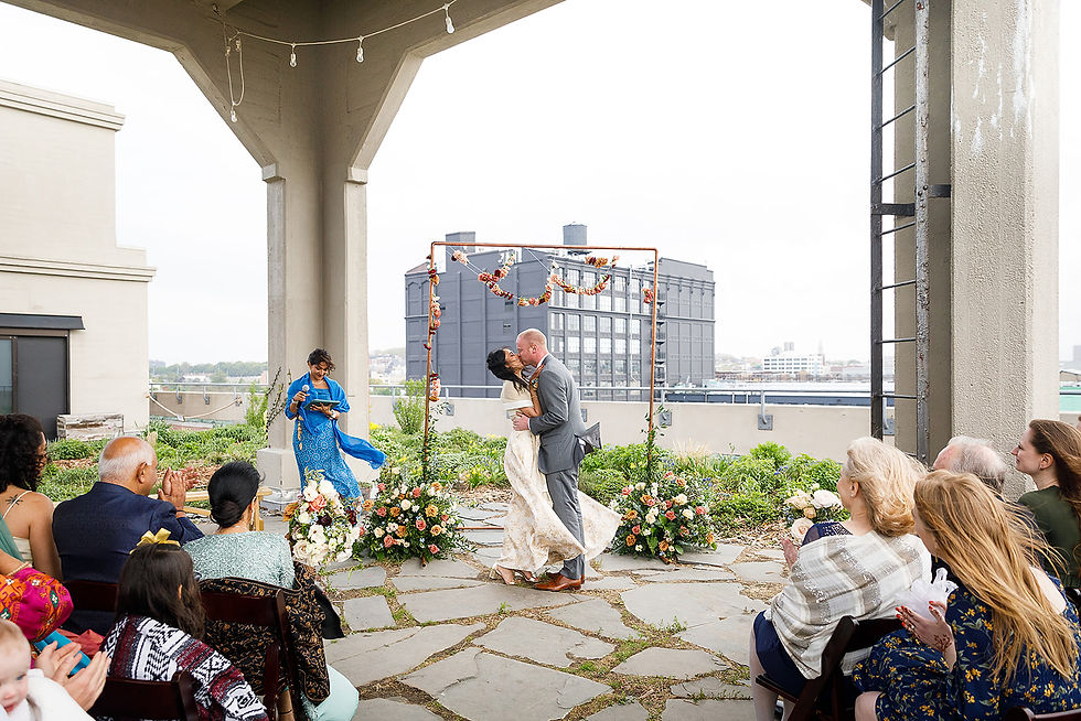 nyc wedding dj new york wedding timeline Bride and groom kiss at a rooftop wedding ceremony, guests applauding under a floral arch with city buildings in the background.