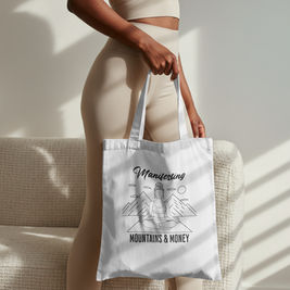 Mountain line-art canvas-like tote bag with the quote "Manifesting Mountains and Money", blending outdoor adventure style with manifestation and empowerment themes.