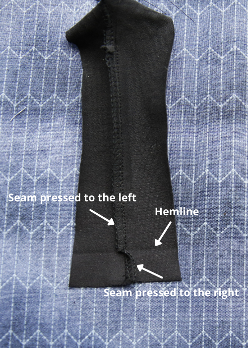 Close up view of an 3 thread overlocked seam in black jersey after snipping the seam at the hemline, showing the seam pressed one way above the hemline and the other way below it to reduce bulk when folding up the hem