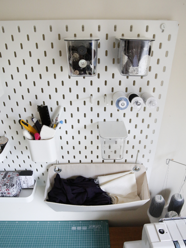 A white Ikea Skadis pegboard with an open container holding sewing tools and 3 clear containers with lids holding buttons and stretch lace, spool holders carrying spools and bobbins and a folder holding fabric