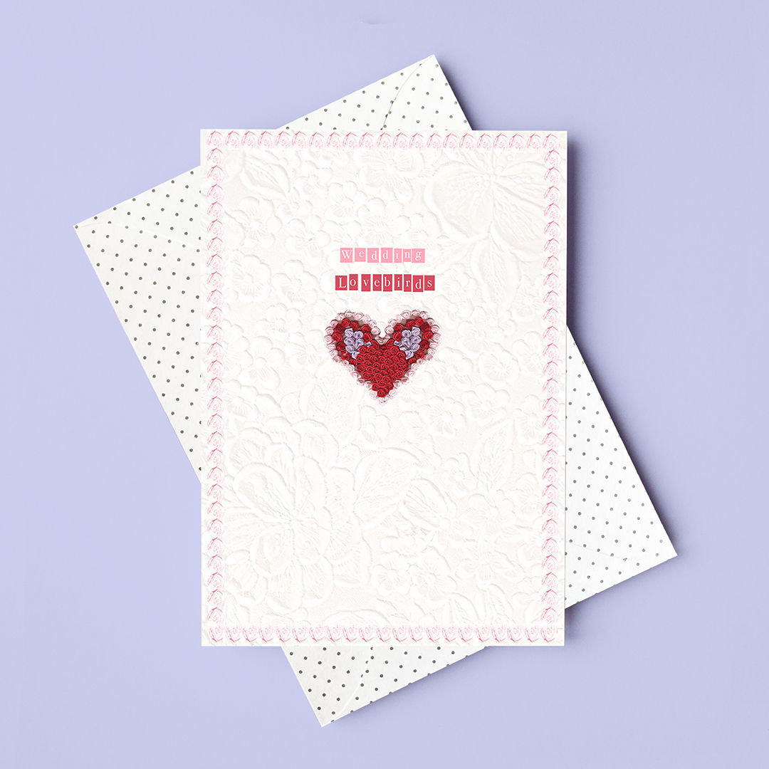 A delicate wedding card showing an illustrated red heart. The card sits on a polka dot envelope and lilac background.
