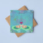 A colourful illustrated greeting card of frogs kissing. The card rests on a square brown envelope and blue background.