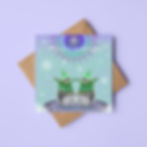 A colourful illustrated greeting card of Christmas Penguins. The card rests on a square brown envelope and lilac background.