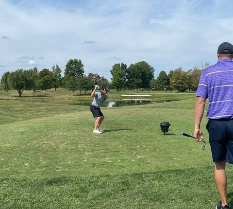 Golfer swings club on a sunny course as another watches.