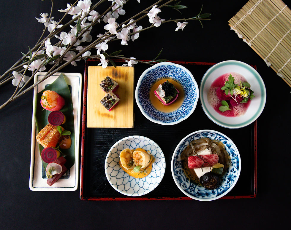 A variety of colorful Japanese dishes artfully arranged on a black tray with a cherry blossom branch and bamboo mat background.