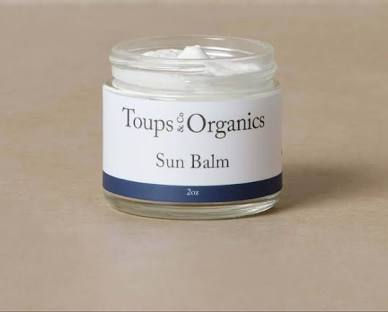 A glass jar labeled "Toups & Organics Sun Balm" with white contents on a beige surface. Label features a blue band and 2oz text.