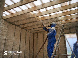 Spray foam insulator applies SPF to the wooden rafters of a ceiling. South Forty Spray Foam