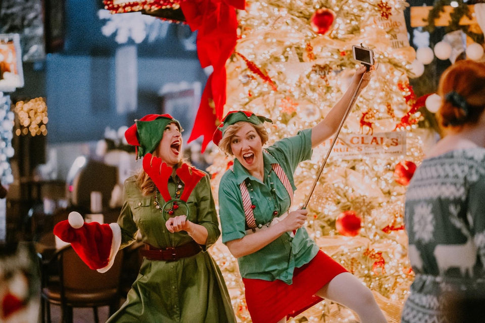 Two women dressed as elves take selfie near Christmas tree.