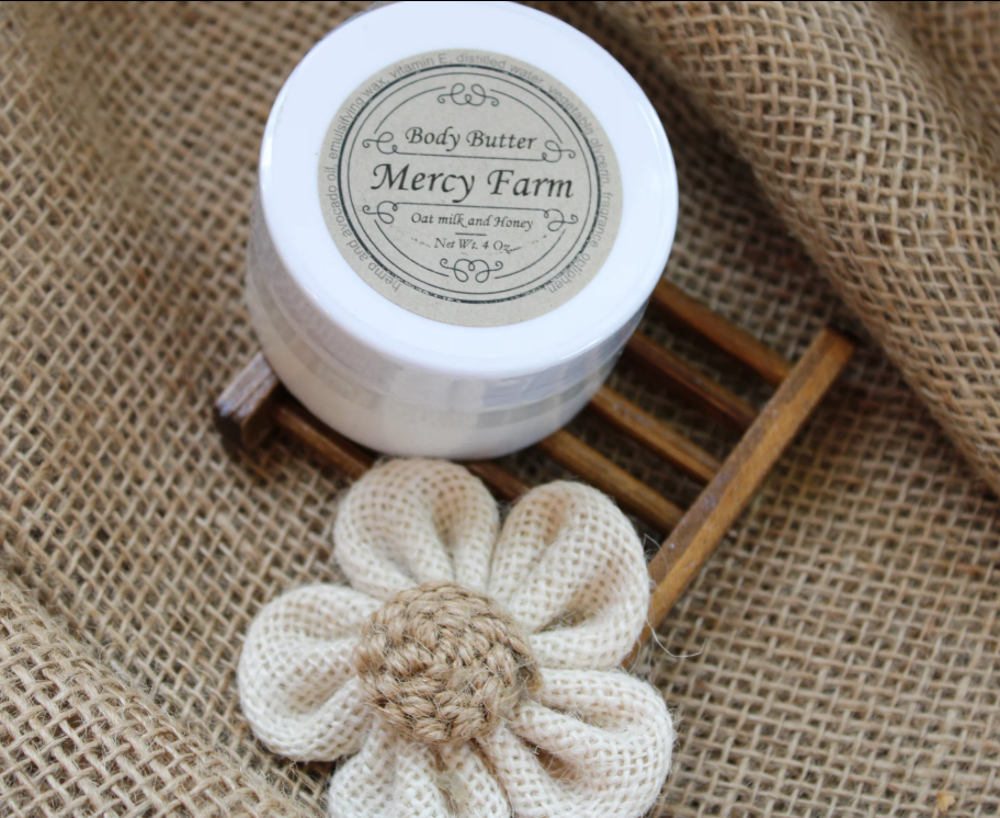 Mercy Farm Oat Milk & Honey Body Butter jar on a small wooden tray with burlap flower on rustic burlap backdrop.