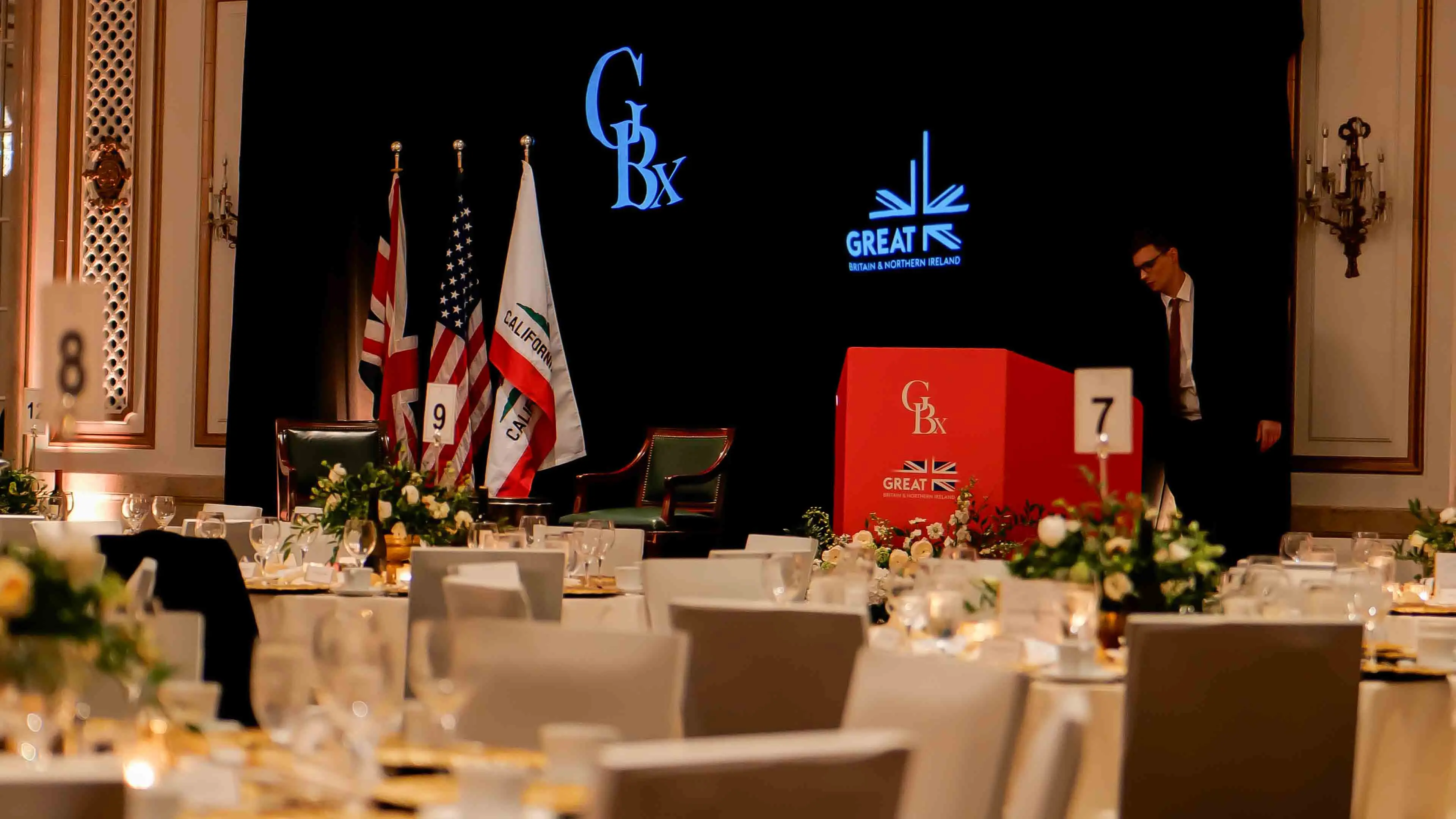 Elegant banquet setup with floral centerpieces and stage at the GREAT GBx Gala 2024 in the Palace Hotel San Francisco, featuring GBx and GREAT Britain branding alongside UK, U.S., and California flags.
