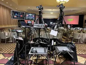 The image shows two cameras on tripods, monitors displaying the live feed, and various other video production equipment. The setup is positioned in a banquet hall with round tables and a stage in the background. Provided by Bidar Video.