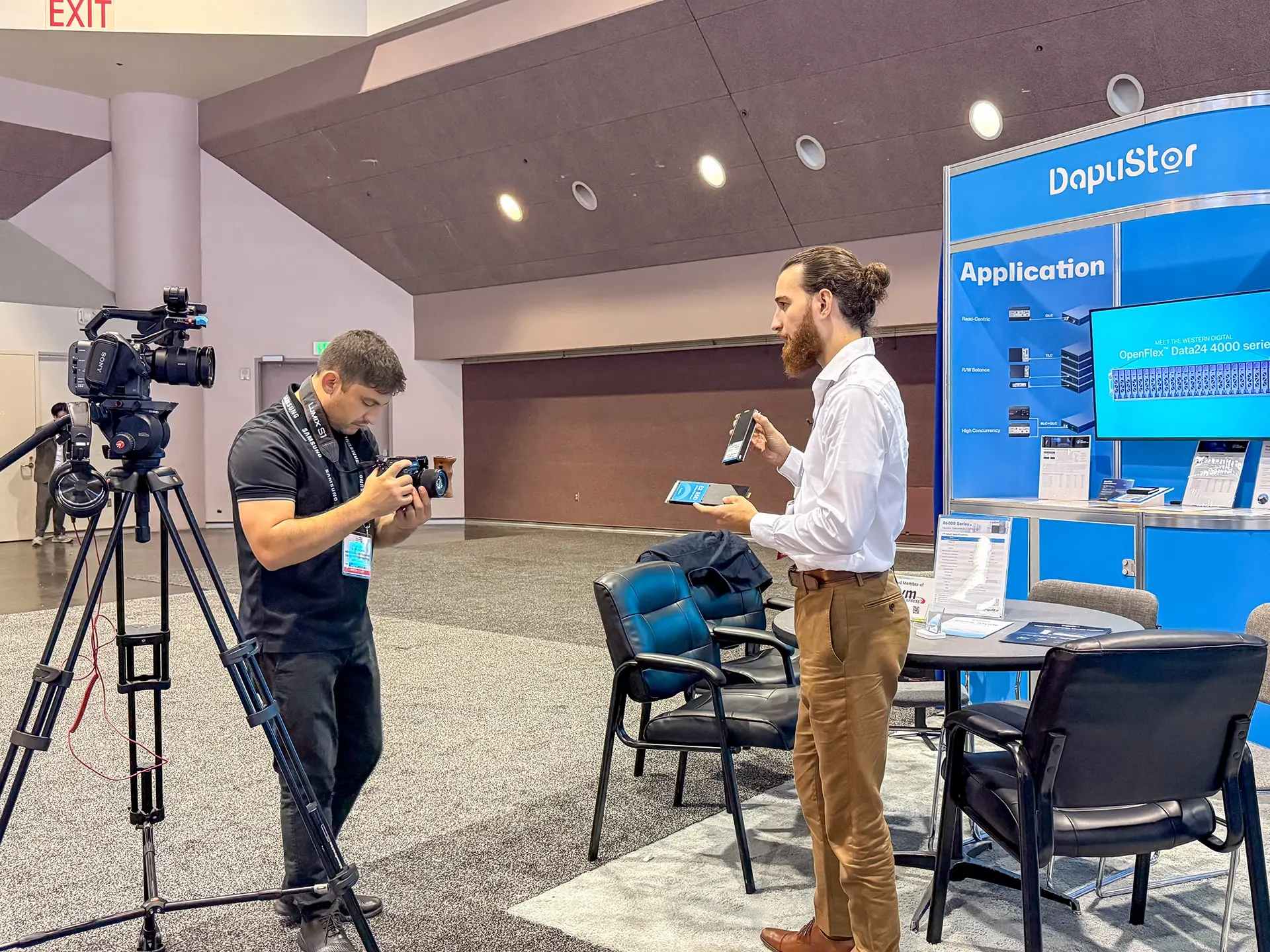 Saboor Bidar filming a Dapustor representative presenting SSD products at FMS 2025 booth in Santa Clara Convention Center, capturing product close-ups and on-camera statements for the trade show sizzle video.