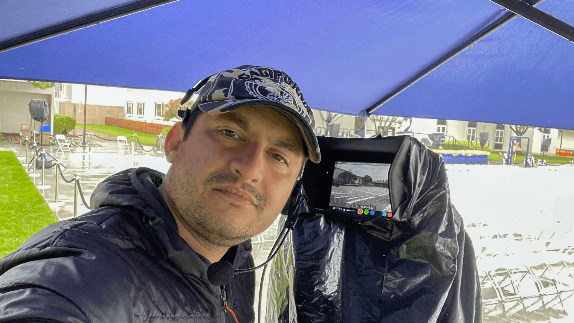 Saboor Bidar under an umbrella moments before an outdoor commencement live stream in windy, rainy weather, with a rain-covered monitor and camera and protected gear from rain and ready to go.
