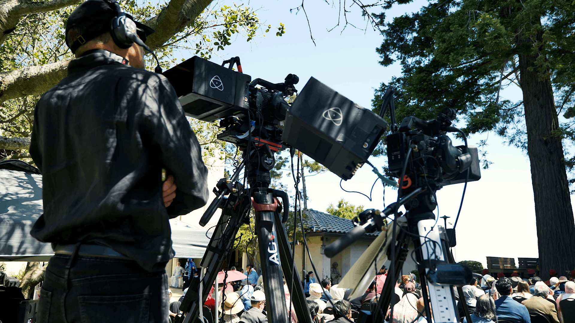 Camera operator wearing headphones beside a tripod-mounted broadcast camera during an outdoor commencement ceremony, with the crowd and trees in the background.