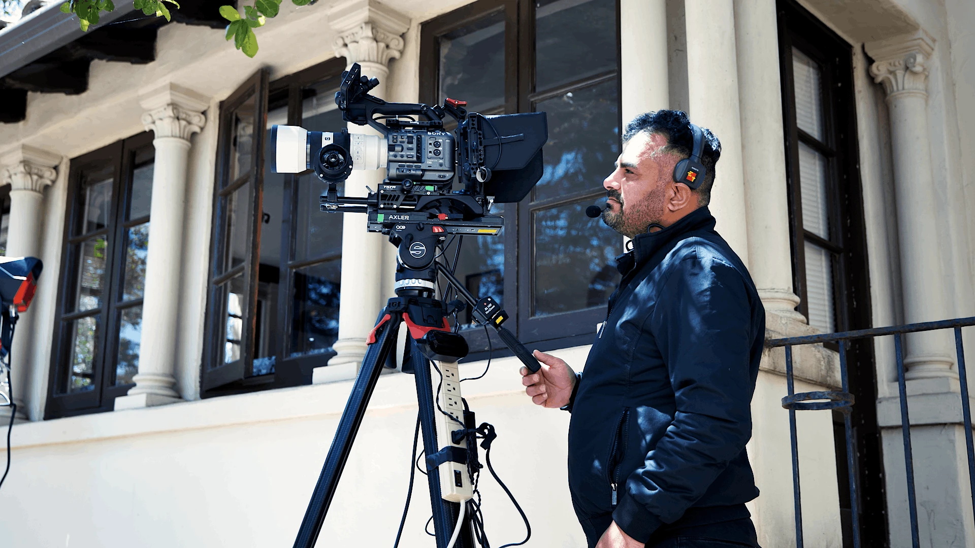 Senior camera operator filming commencement coverage on a tripod-mounted Sony FX6 camera outside a campus building, wearing headphones for audio monitoring