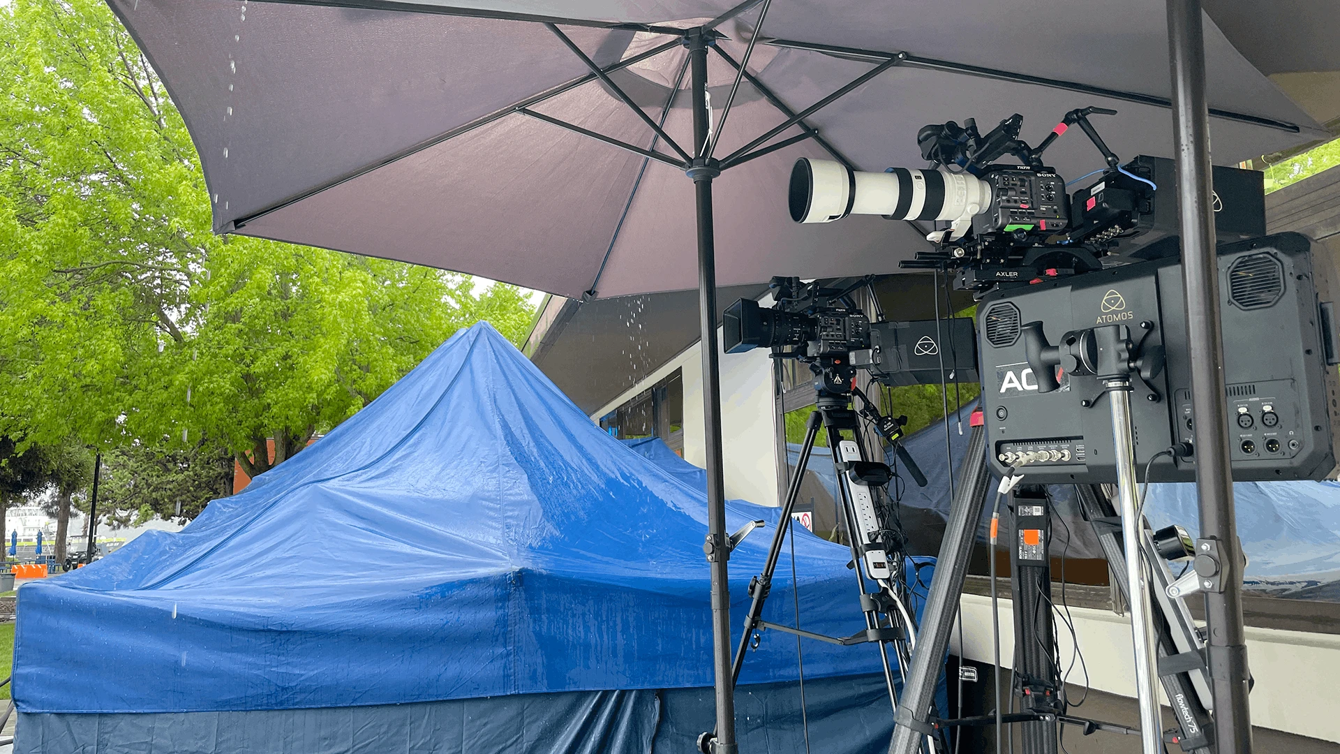 Tripod-mounted cameras with rain cover under an umbrella during an outdoor commencement live stream in overcast, light rain, with pop-up tents protecting the production area from rain and wind..