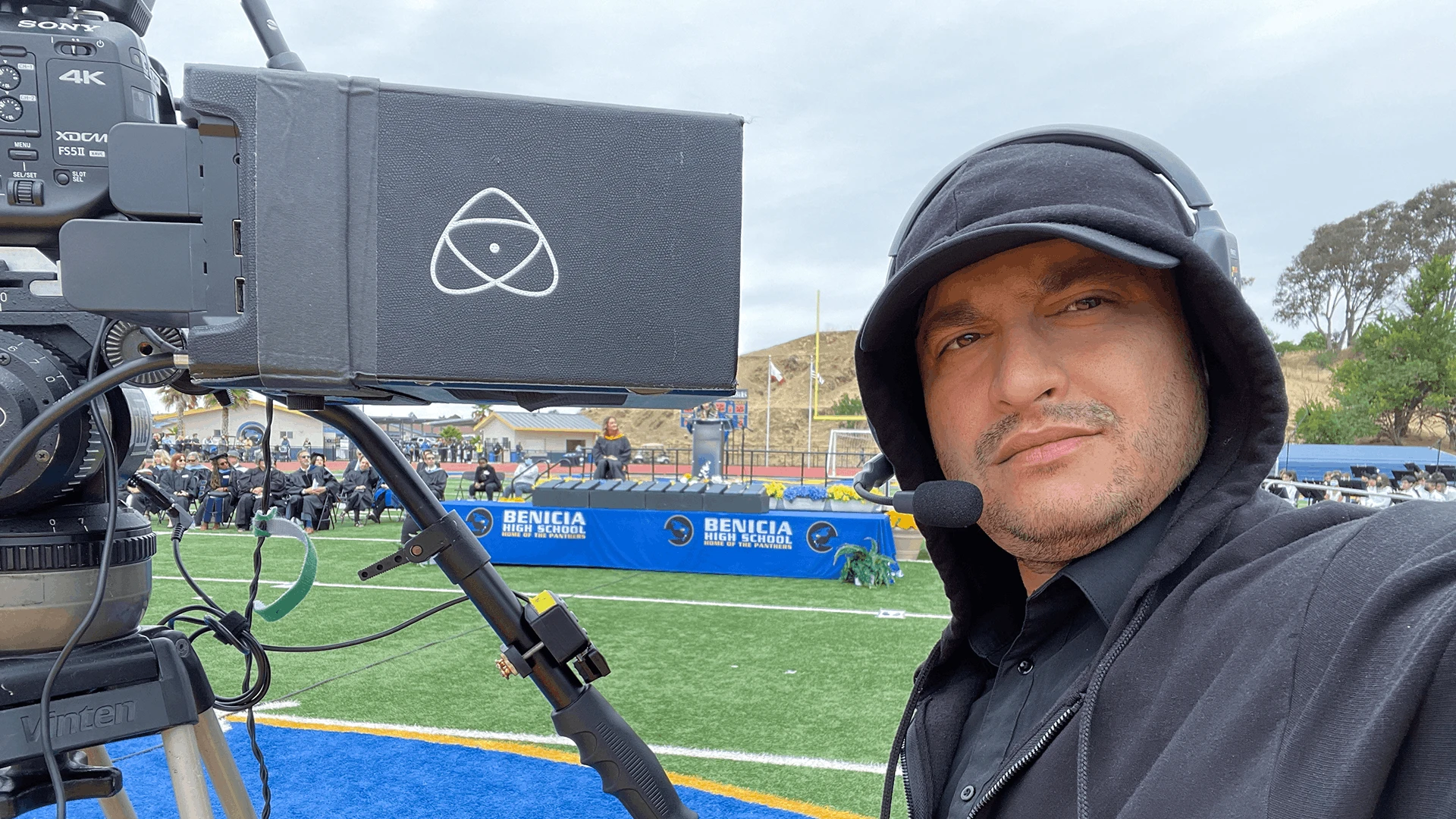 Saboor Bidar wearing a headset beside a tripod-mounted broadcast camera during an outdoor commencement live stream in overcast, light rain, with the ceremony field and audience and stage in the background in a rainy moment.
