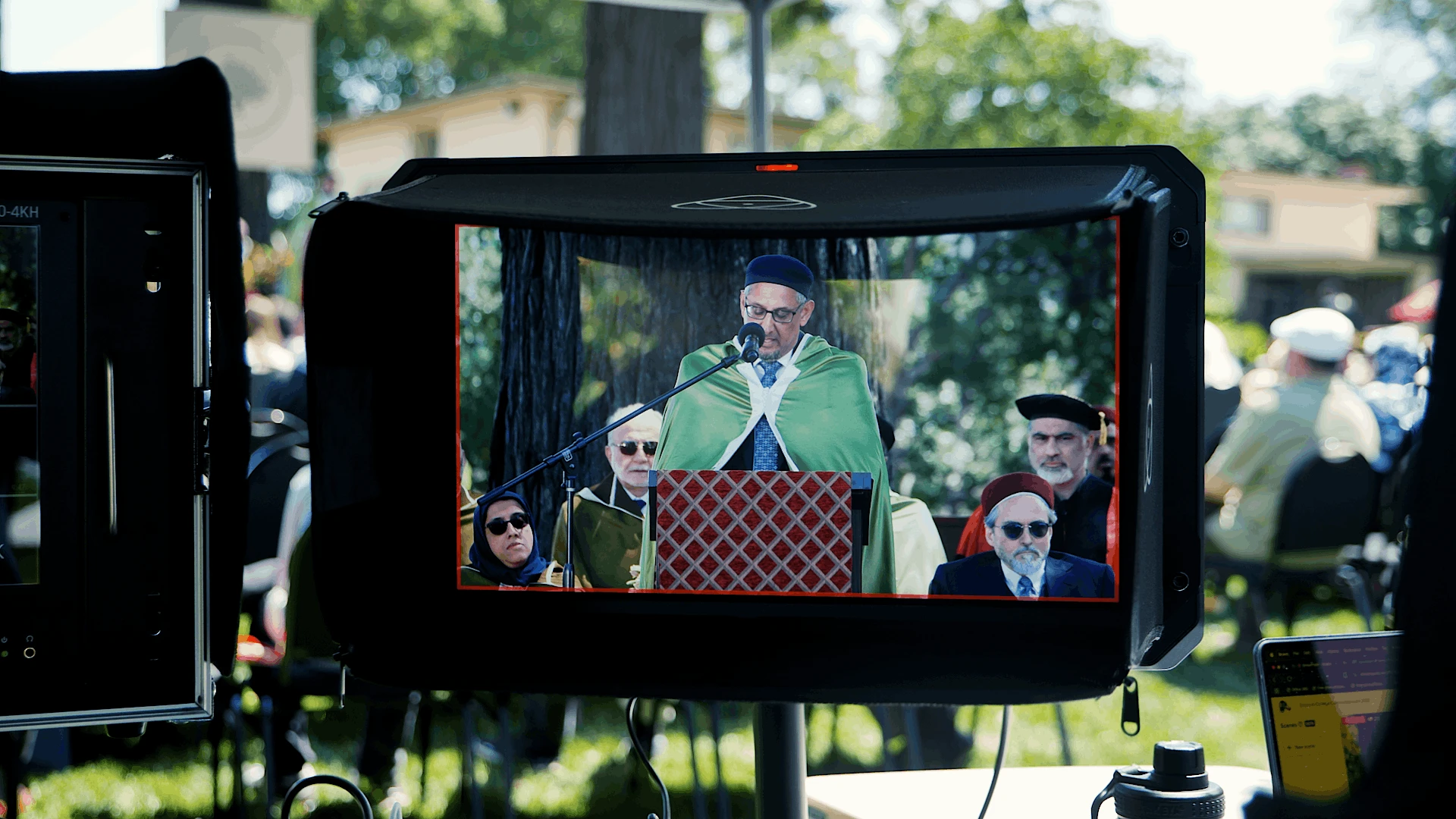 On-camera monitor showing a speaker at the podium during an outdoor commencement ceremony, framed for the live stream with faculty seated behind.
