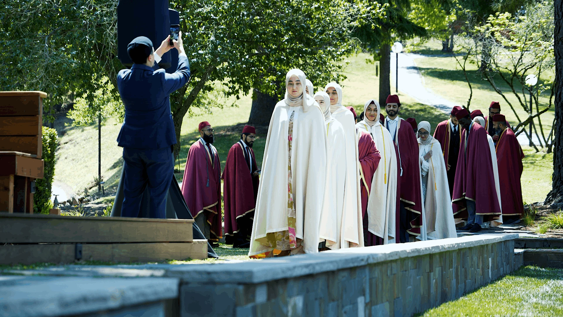 Faculty and graduates in ceremonial robes walking in procession outdoors while a camera operator films from a raised platform.