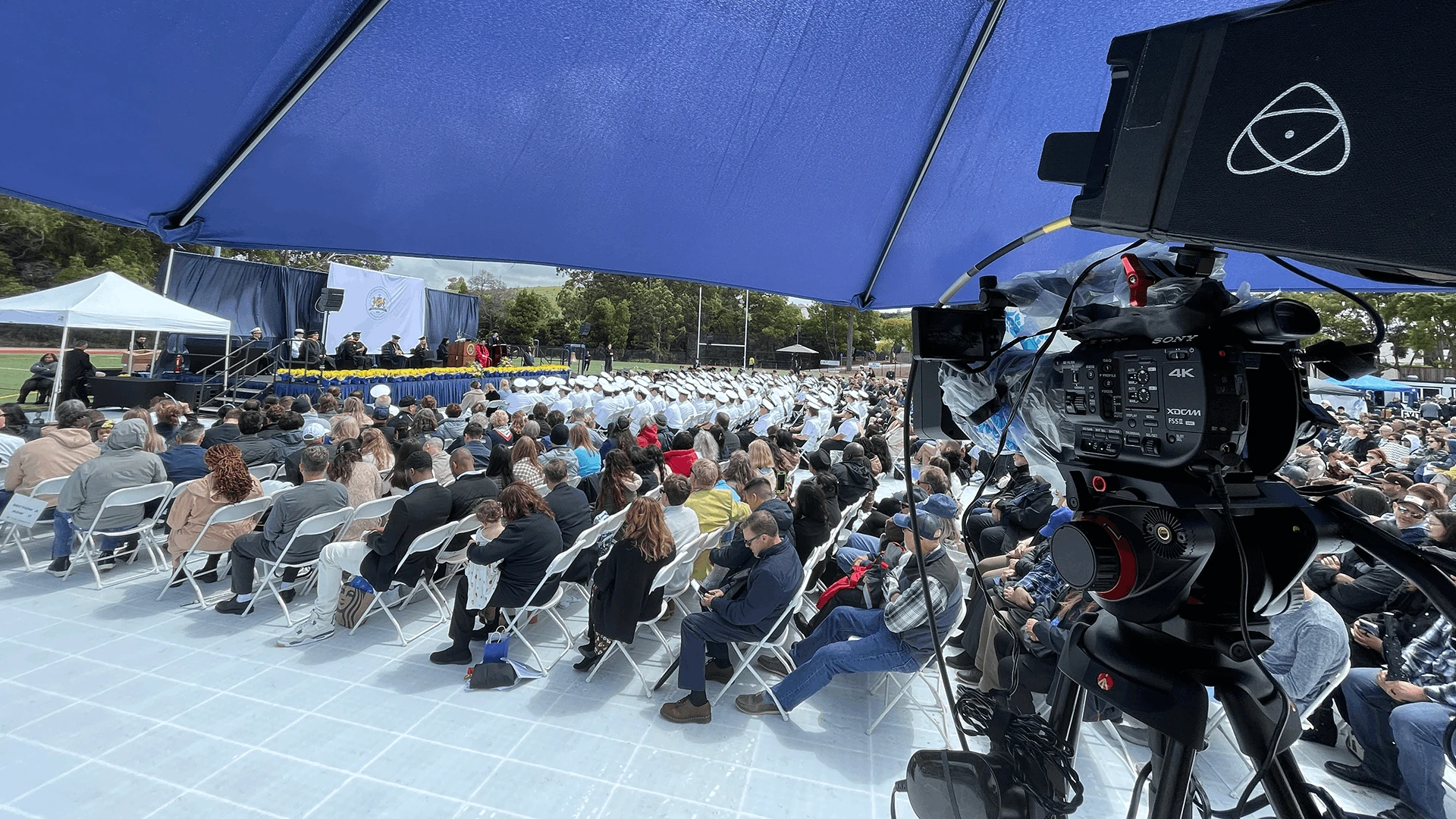 Broadcast camera filming an outdoor commencement ceremony in overcast, light rain, sheltered under an umbrella with the seated audience visible in the background while light rain..