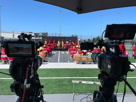 Two professional video cameras on tripods filming an outdoor high school graduation ceremony on a football field, with graduates in caps and gowns crossing the stage.