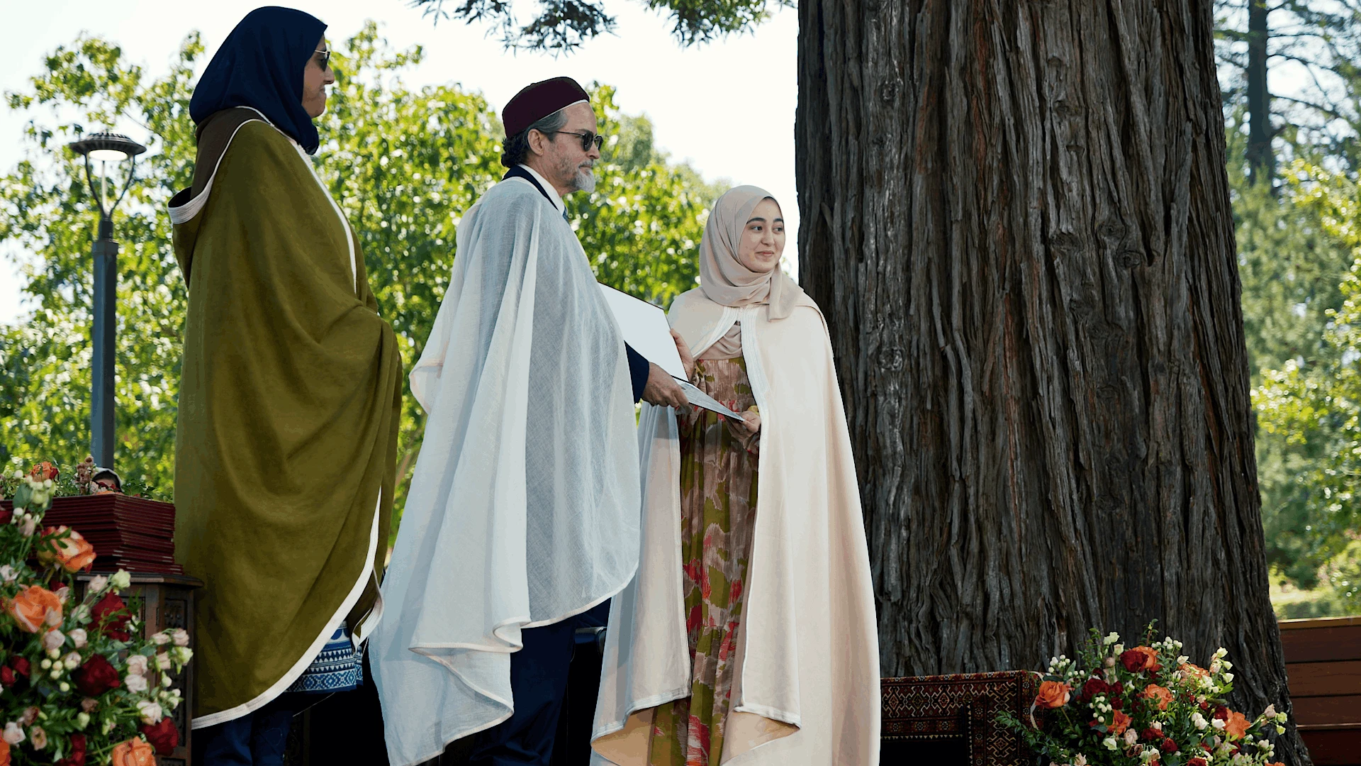 Faculty and graduates in ceremonial robes standing on an outdoor commencement stage near a large tree and floral arrangements.
