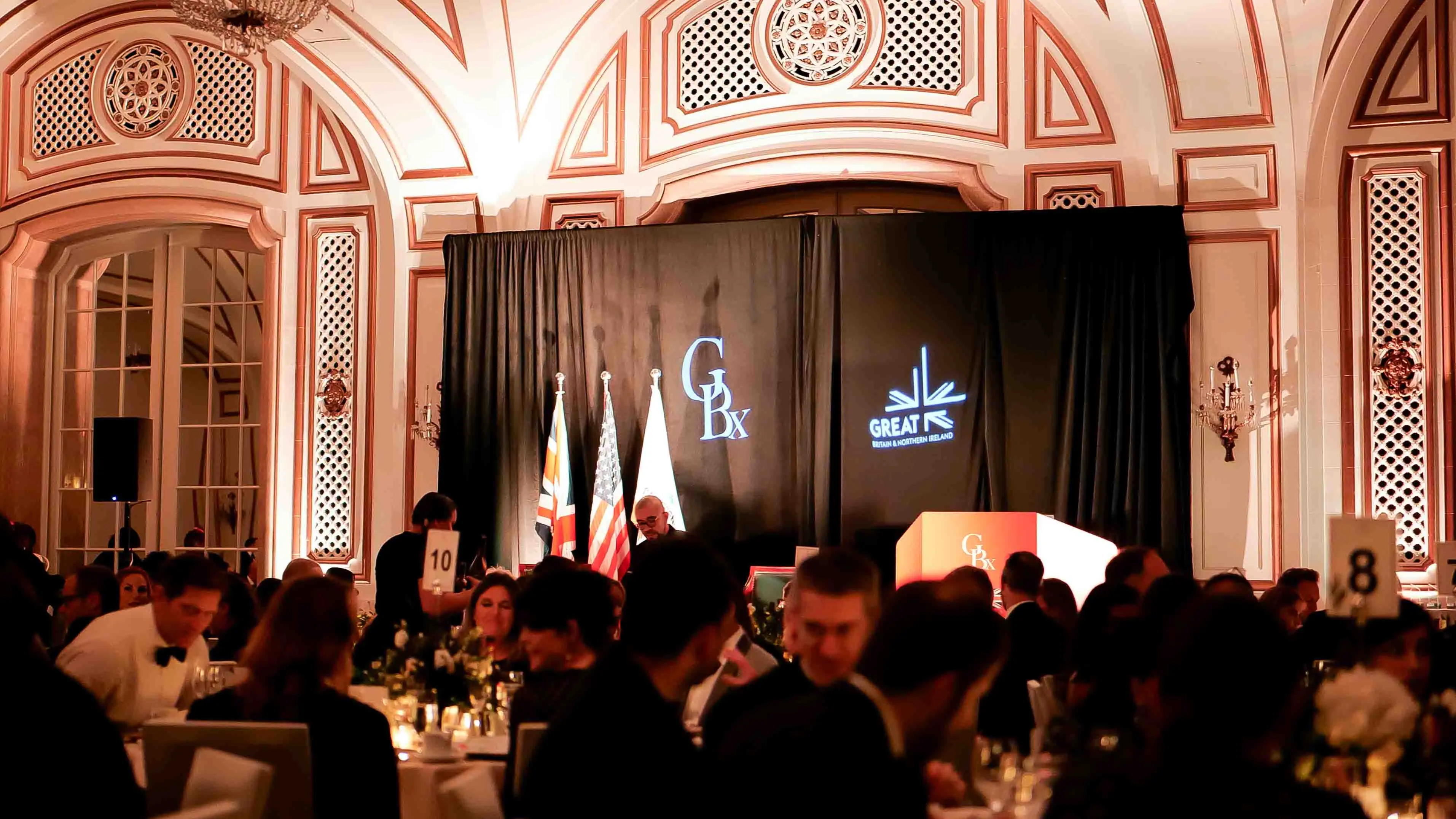Guests dining at the GREAT GBx Gala 2024 inside the grand ballroom of the Palace Hotel San Francisco, with stage lighting, international flags, and elegant decor highlighting the UK–U.S. partnership.