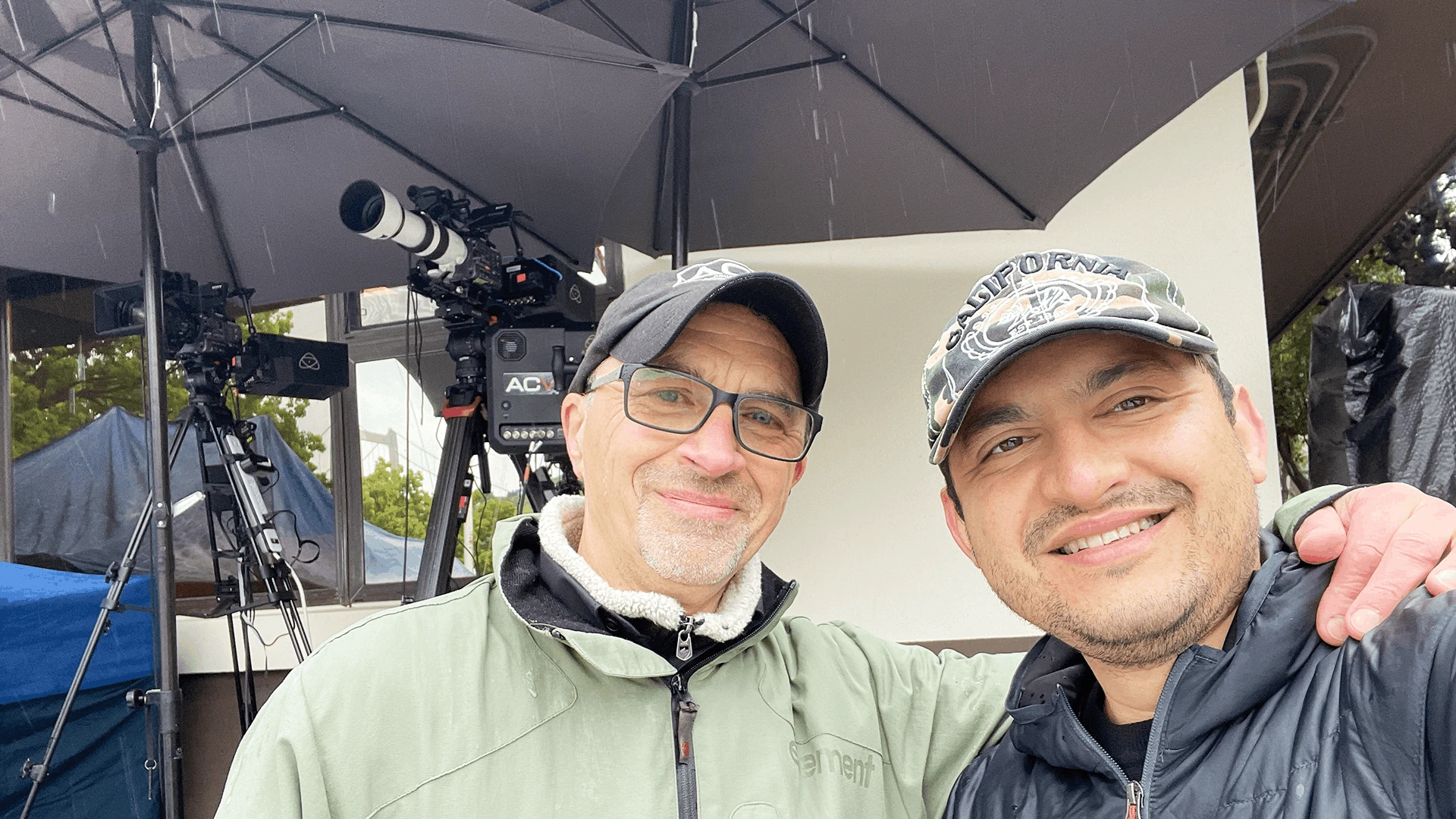 Saboor Bidar and Alan Cash smiling under umbrellas after a successful outdoor graduation live stream in overcast, light rain, with a camera setup behind them after successfully completing the live streama in a challenging weather condition.