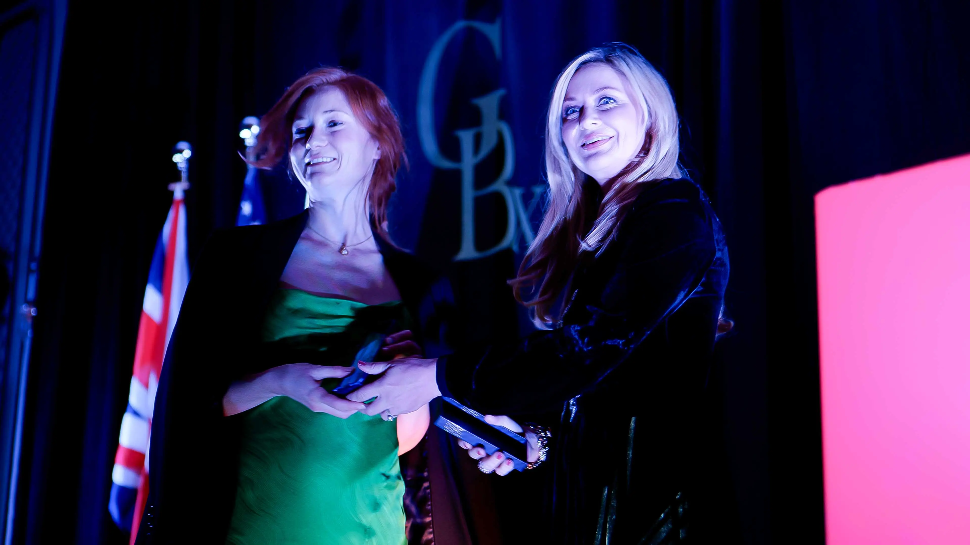 Two women on stage smiling and holding an award during the GREAT GBx Gala 2024 at the Palace Hotel in San Francisco, with GBx branding and UK flag in the background.