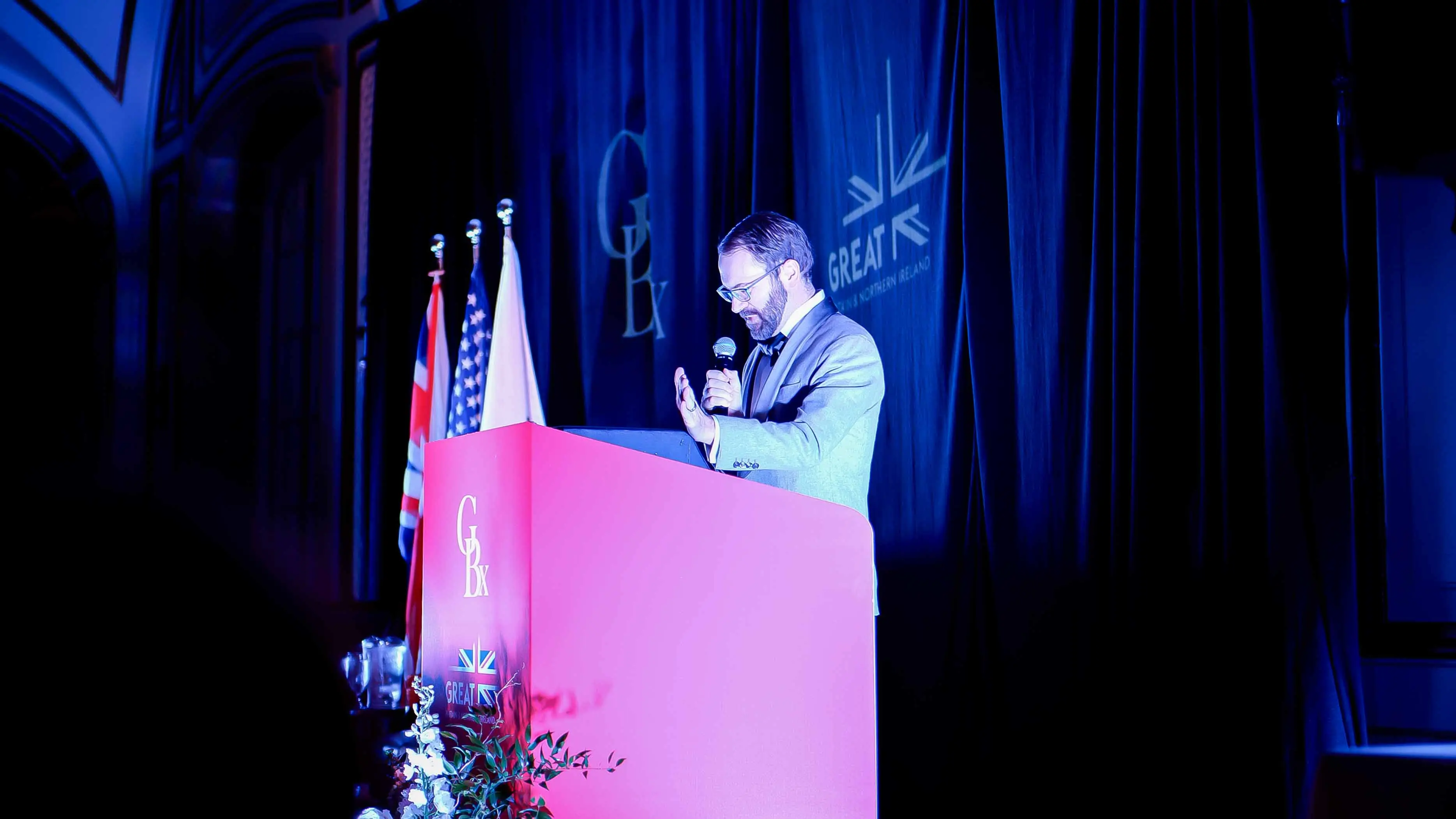 Speaker delivering remarks from the podium during the GREAT GBx Gala 2024 at the Palace Hotel in San Francisco, with GBx and GREAT branding and international flags in the background.