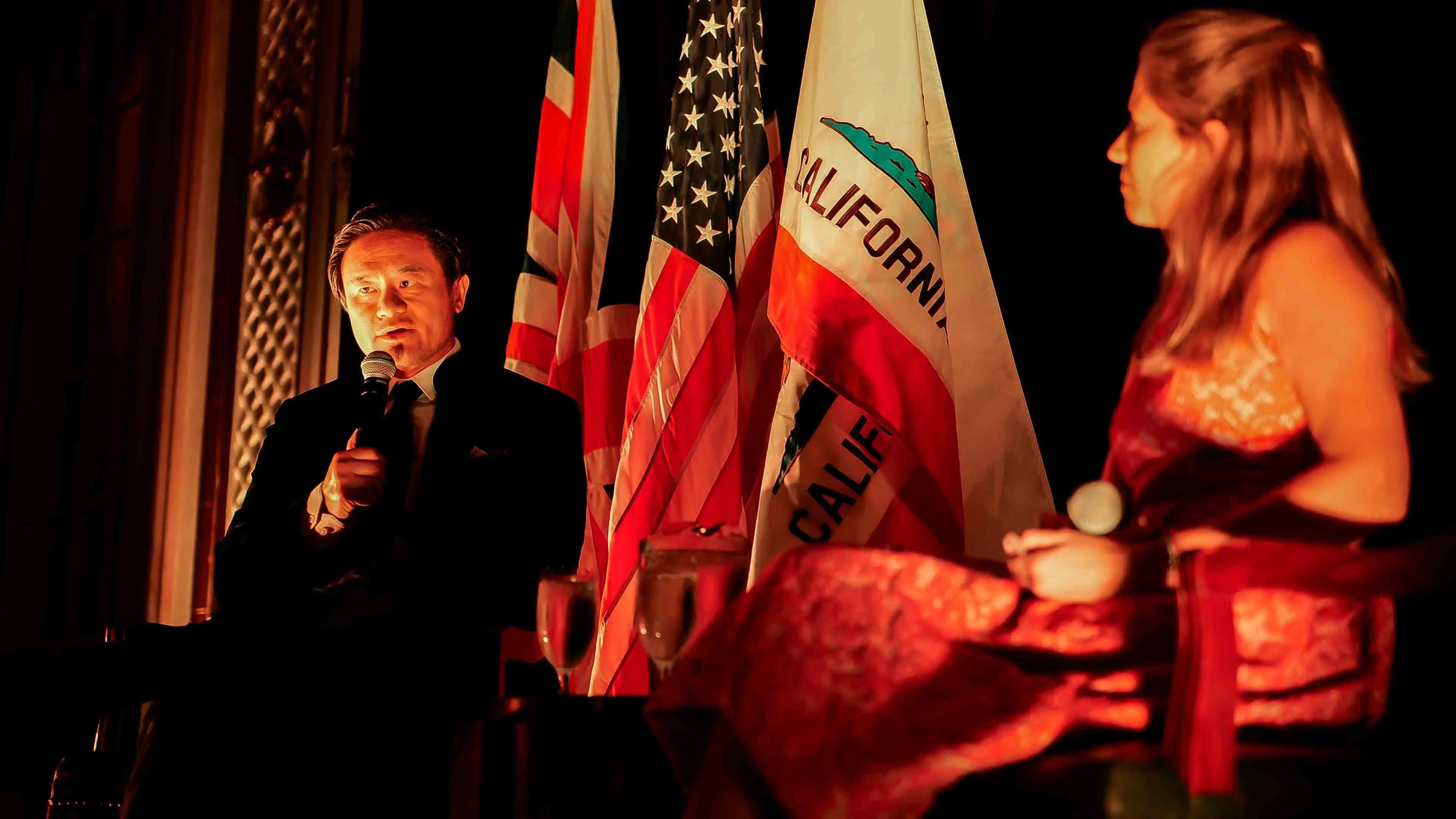 Speakers engaged in a fireside chat during the GREAT GBx Gala 2024 at the Palace Hotel in San Francisco, seated in front of the UK, U.S., and California state flags under warm stage lighting.