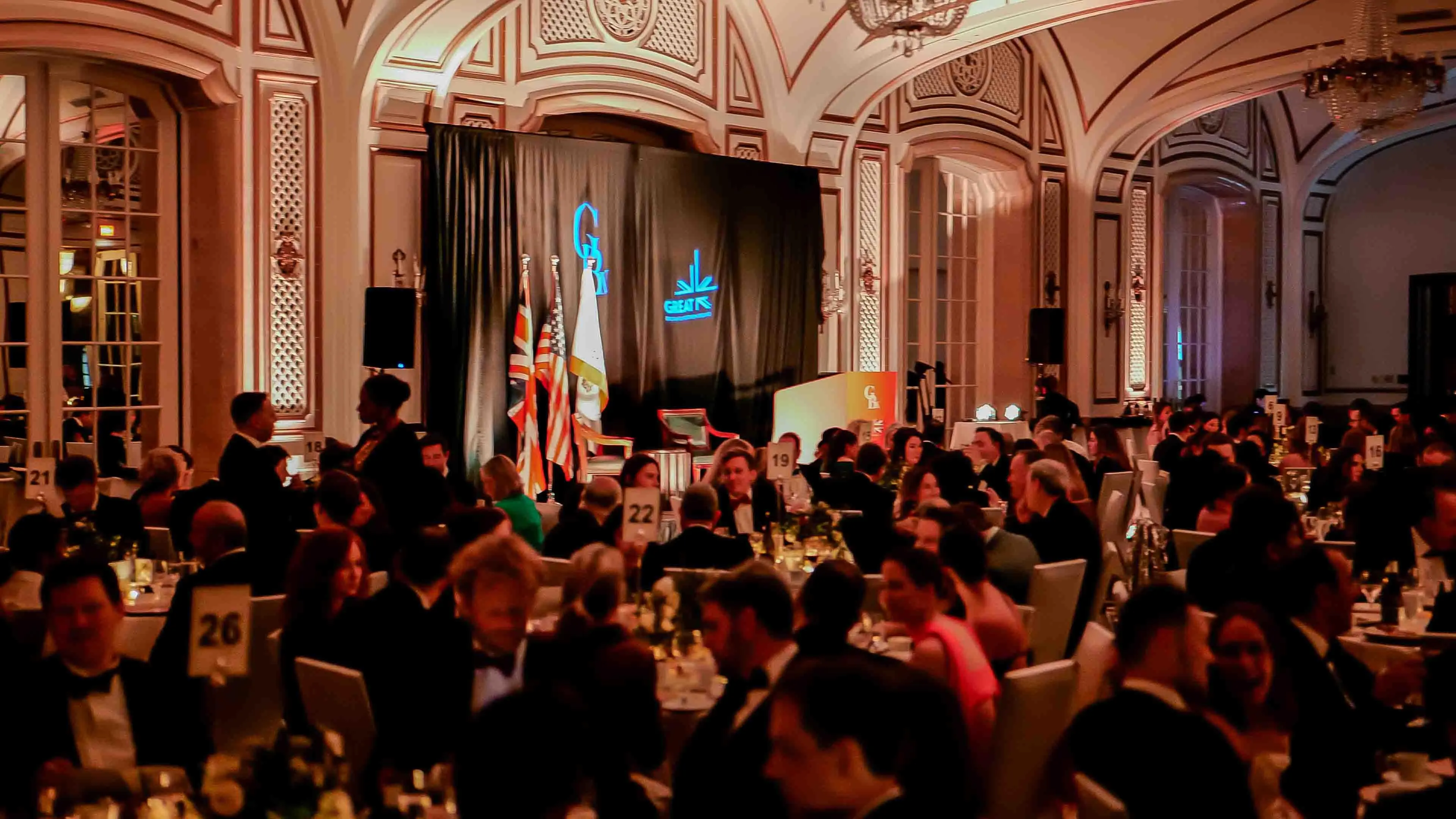 Wide view of the ballroom at the Palace Hotel in San Francisco during the GREAT GBx Gala 2024, filled with formally dressed guests seated at candlelit tables in front of a branded stage with international flags.