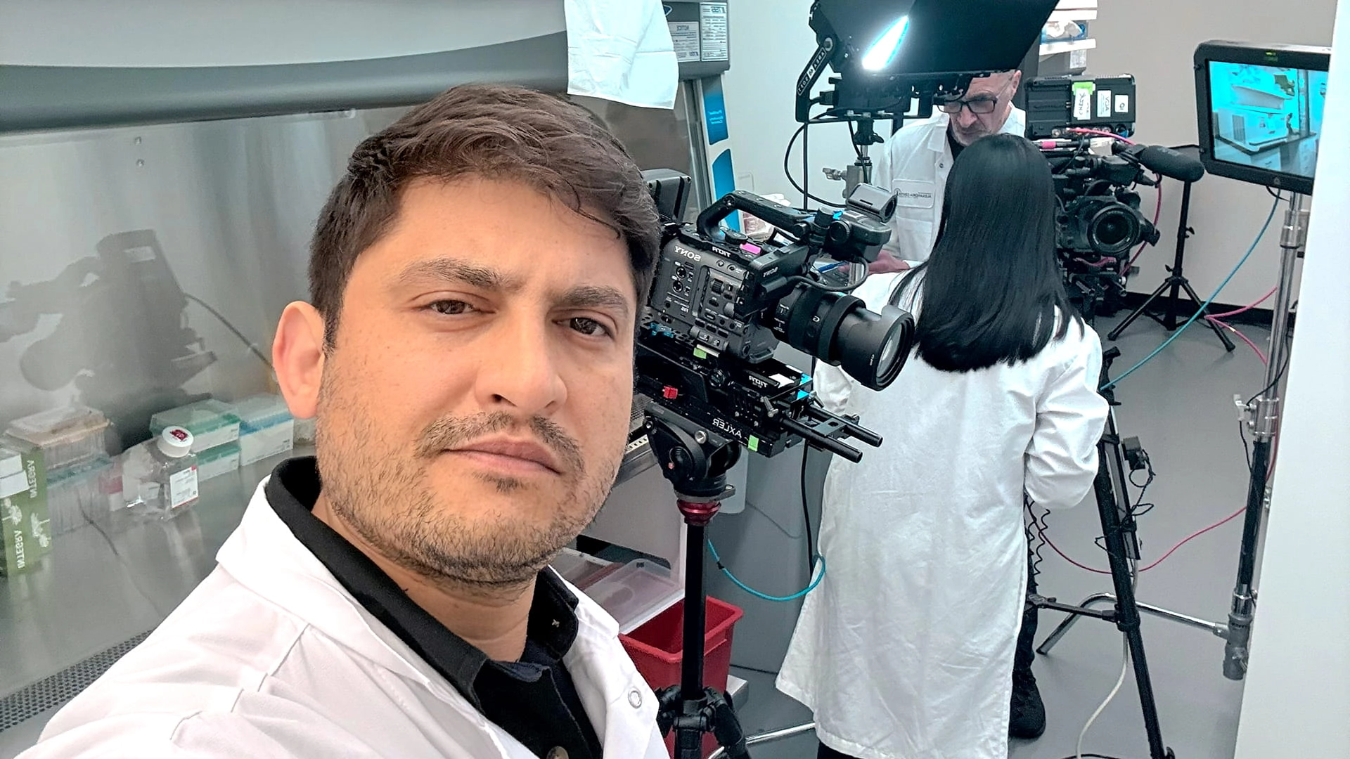 Saboor Bidar in a biotech lab during a training video shoot, with cameras, lighting, monitors, and on-camera procedure filming visible in the background.