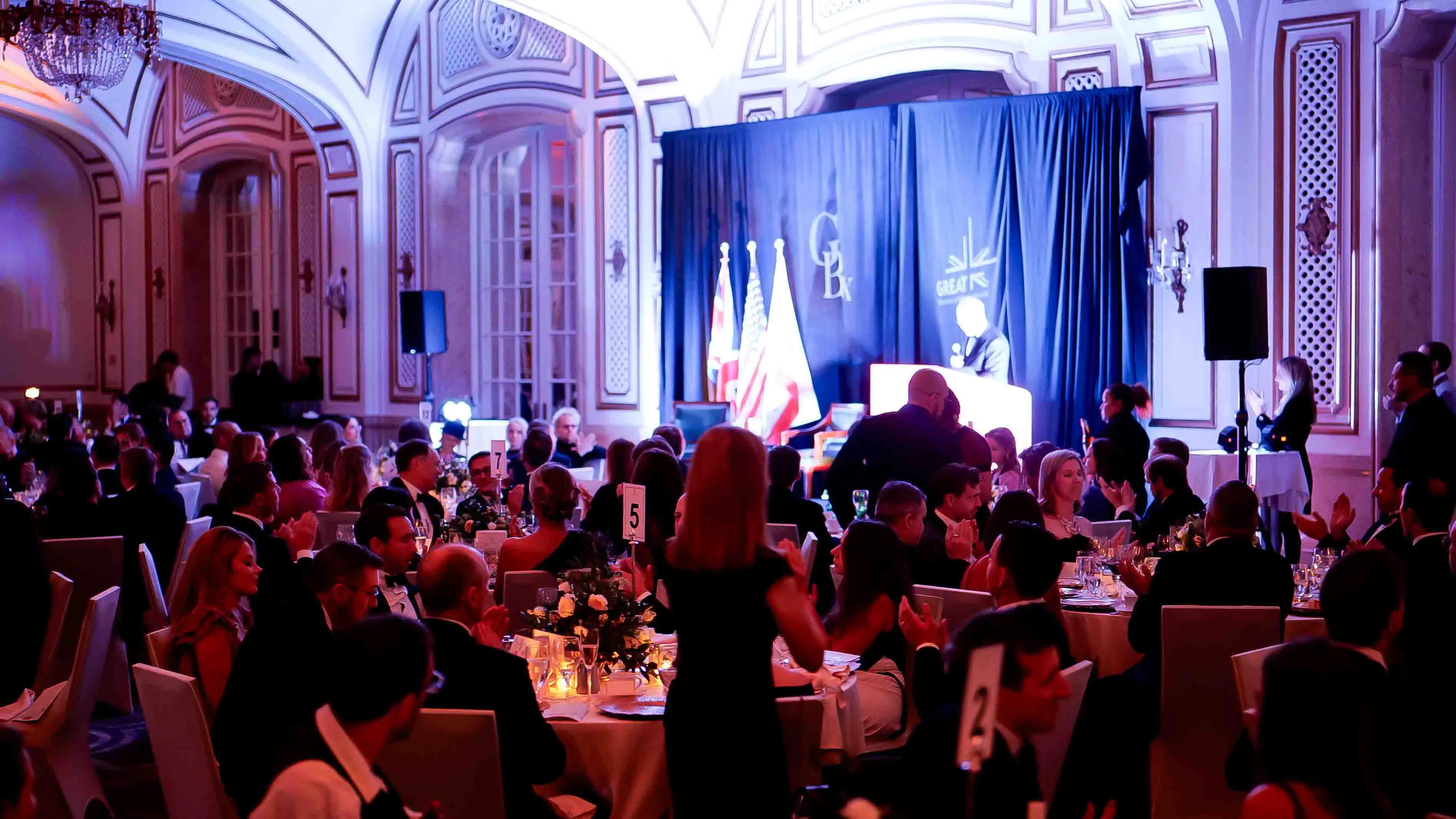 Elegant ballroom filled with attendees as a speaker presents on stage at the GREAT GBx Gala 2024 in San Francisco, with international flags and ambient lighting enhancing the event atmosphere.