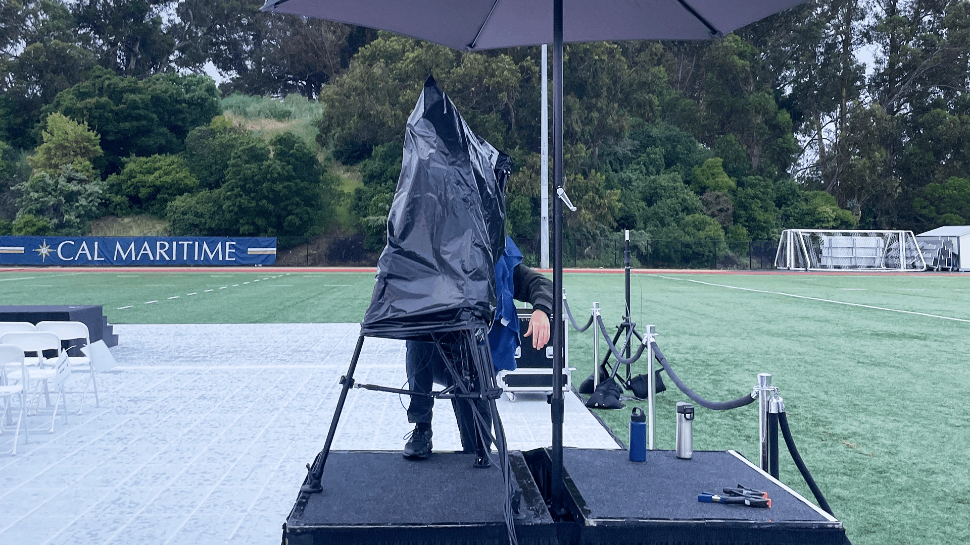 Cameras set up on an outdoor field the day before commencement, covered with weather-protection plastic and secured under an umbrella overnight to be ready for the next day’s live stream from rain..