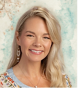 A smiling woman with long blonde hair and dangling earrings poses in front of a light, textured backdrop. She wears a floral-patterned top.