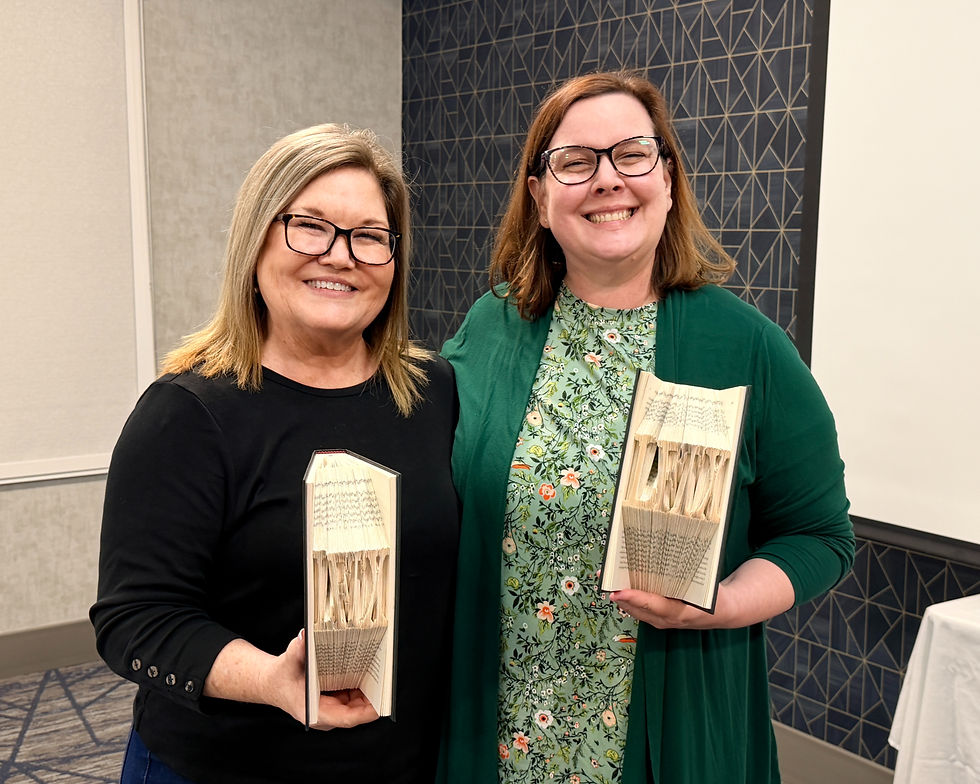 Two smiling women in an office hold books with fanned pages, standing before patterned walls and a table with markers.